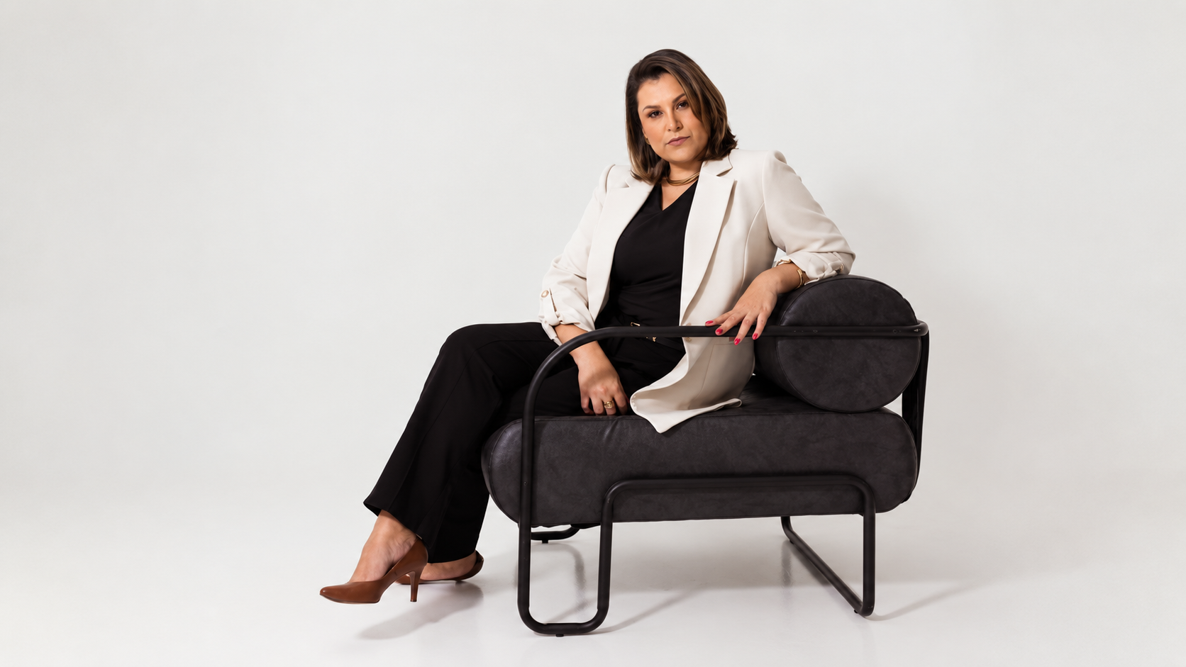 Karin Rost sentada em uma poltrona de couro escuro, blazer claro sobre roupa preta, diante de um fundo branco de estúdio.
